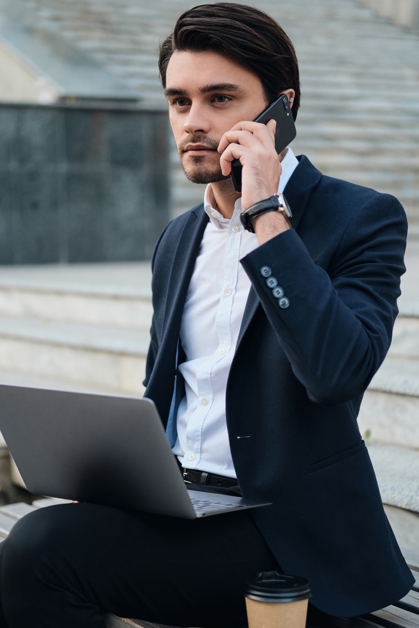 Young businessman in suit sitting on bench with laptop dreamily looking aside talking on cellphone